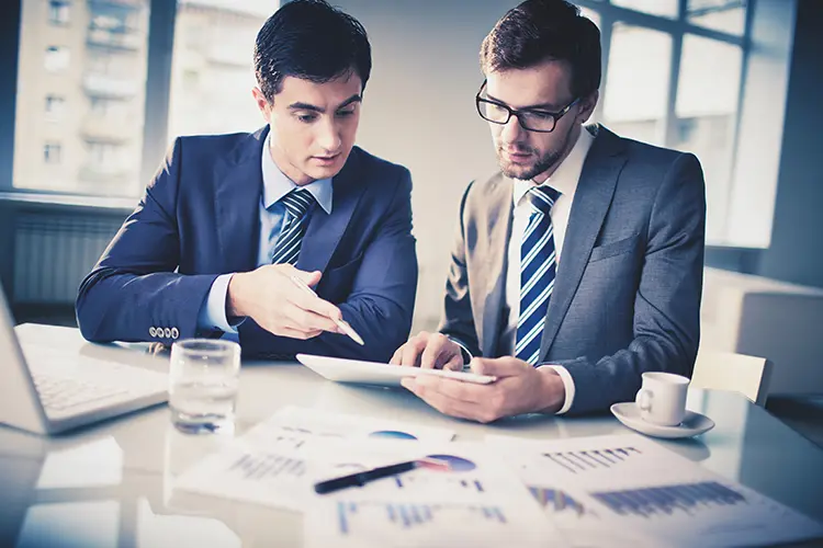 Two professionals discussing work with a tablet in a modern office with large windows.