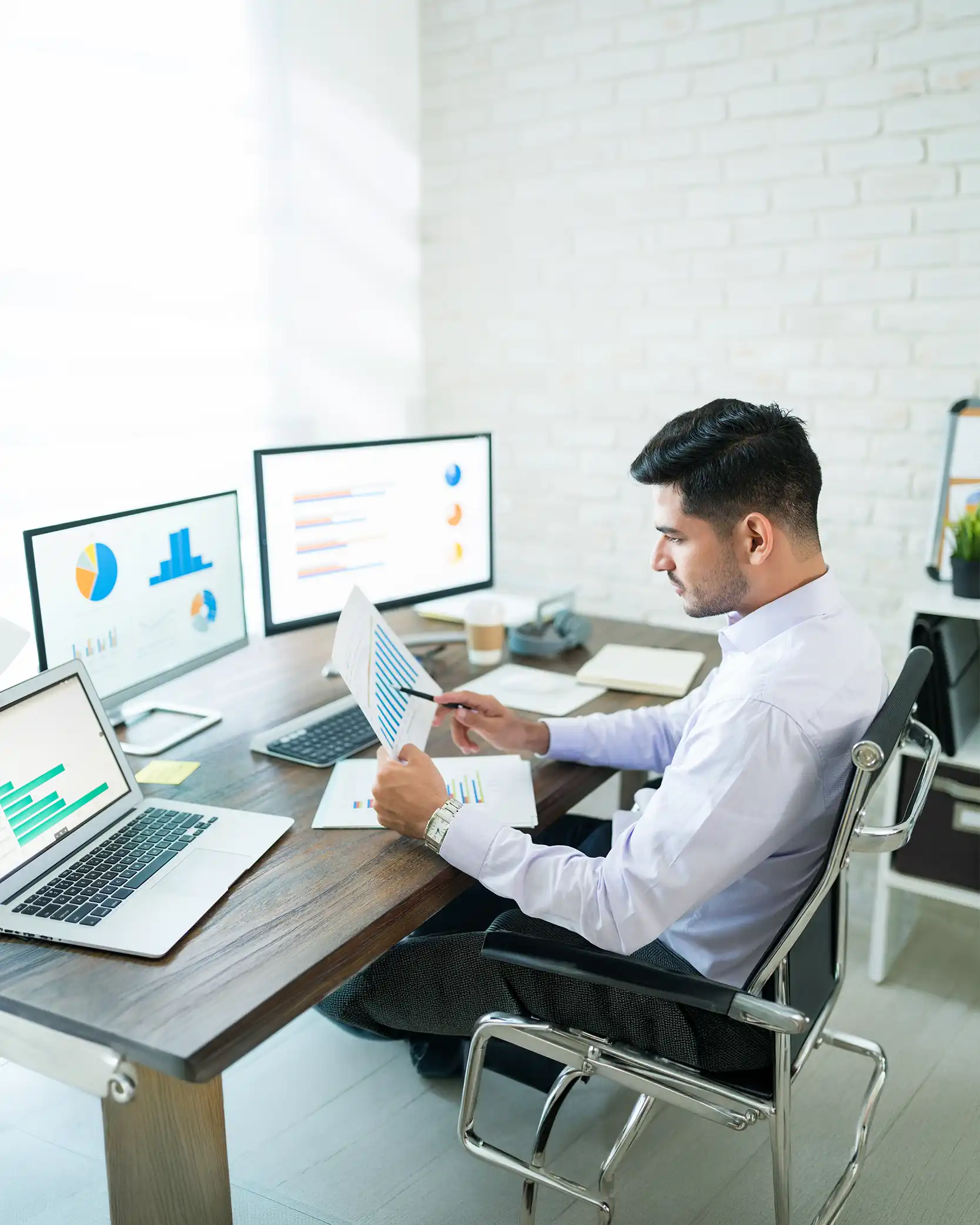 A man working at a desk with multiple monitors and analyzing charts, in a modern office setting.