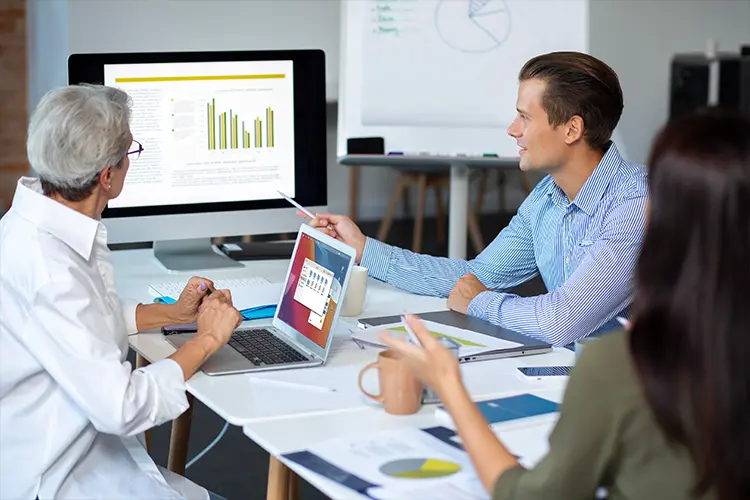 Team of professionals discussing data and charts on computer screens in a business meeting or collaborative office setting.