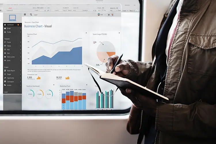 Person in a jacket taking notes while analyzing business charts on a tablet, with digital dashboards displayed on a large screen behind.