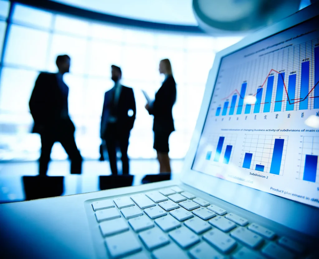 Laptop screen displaying charts and graphs, with three business professionals in the background discussing in a modern office.
