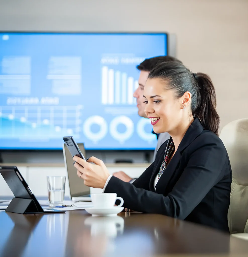 A woman in business attire smiling and looking at her phone during a meeting, with a man in the background and a large digital screen displaying charts and graphs.