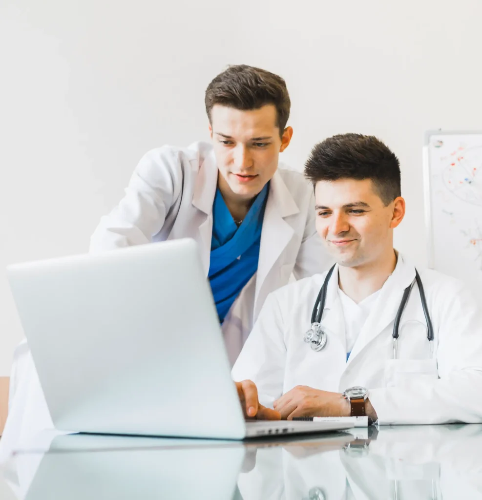 Two male doctors in white coats, one with a stethoscope, looking at a laptop screen together in a medical office or hospital setting.