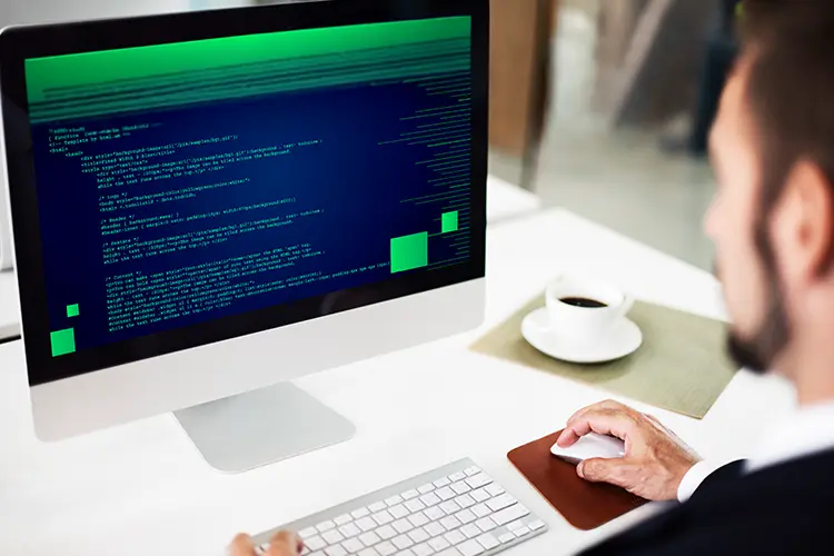 A person working on a computer with green and blue code on the screen, sitting at a desk with a cup of coffee nearby.