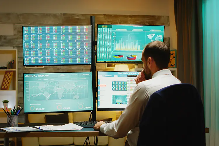 Man working at a desk with four computer monitors displaying charts, graphs, and data in a modern office setting.