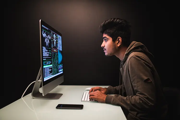 A young man in a hoodie working intently on a computer with code and data displayed on the screen, in a dark room.