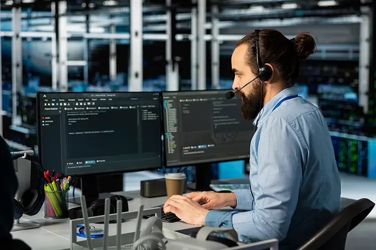 A man with a headset works at a computer with dual monitors in a server room filled with racks of servers.
