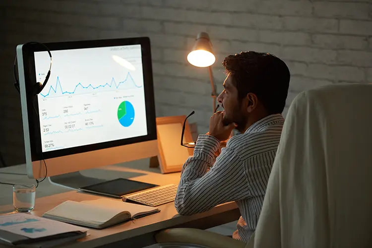 A man in a striped shirt works at a desk with a computer displaying graphs and charts, illuminated by a desk lamp in a modern office.