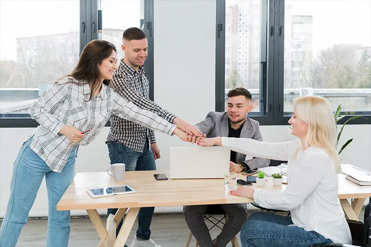 Group of diverse colleagues in an office, with three people reaching out for a handshake over a laptop, symbolizing teamwork and collaboration.
