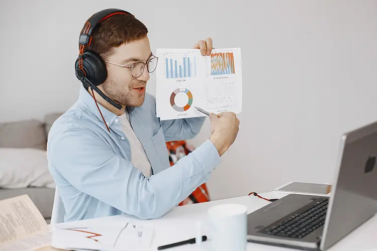 Man with headset showing data charts on paper during a virtual meeting or remote work, with a laptop and documents on the table.