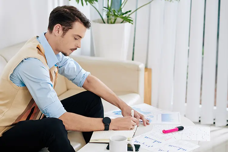 A man sitting on a sofa, working with documents and charts on a table, with a pen in hand, in a bright room with large windows and indoor plants.
