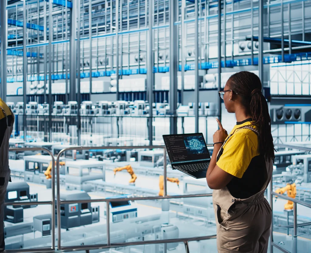 Woman in a yellow shirt using a laptop and pointing in a high-tech warehouse with robotic arms and machinery.