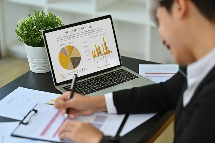 A person in a business suit analyzes monthly budget data on a laptop screen with pie charts and bar graphs, taking notes on printed documents at a desk.