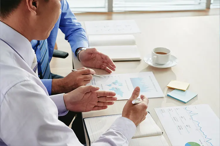 Two professionals discuss data and charts at a desk with documents, a cup of coffee, and sticky notes in a bright office.