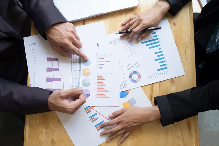 Two people in business suits analyze and discuss colorful charts and graphs on papers spread out on a wooden table.