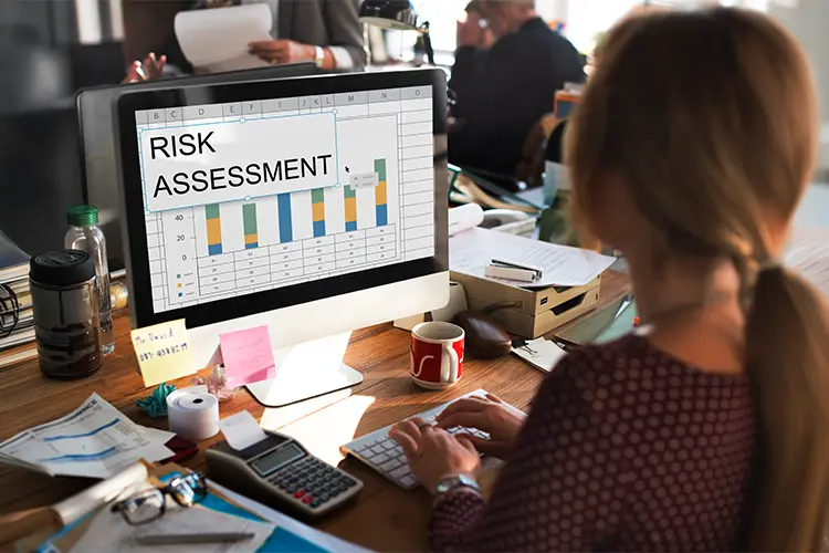 A woman working on a computer with a risk assessment chart on the screen, surrounded by office supplies and colleagues in a busy office environment.