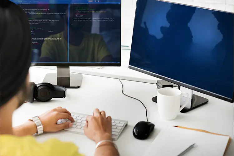 A person working at a desk with dual monitors, coding on one screen, a mug, headphones, and a notebook, in a modern office environment.
