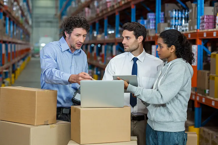 Three colleagues discussing inventory or logistics in a warehouse, with shelves filled with boxes and products in the background.