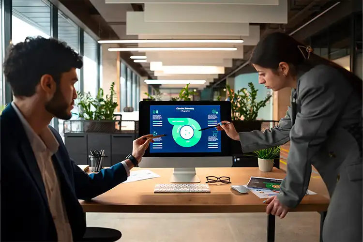 Two professionals in business attire discuss data and charts displayed on a computer monitor in a modern office, with plants and large windows in the background.